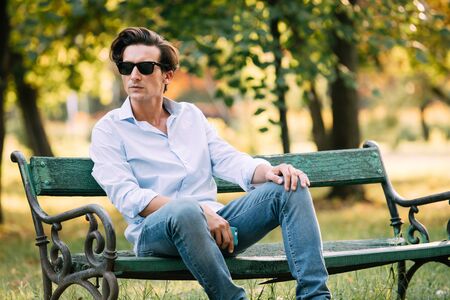 attractive man in blue shirt sitting alone on the bench in park with cellphone and sunglassesの写真素材