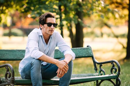 attractive man in blue shirt sitting alone on the bench in park with cellphone and sunglassesの写真素材