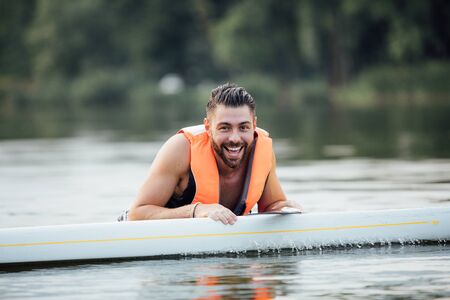 handsome wet man standing in water and smiling on a paddleboard surrounded by treesの写真素材