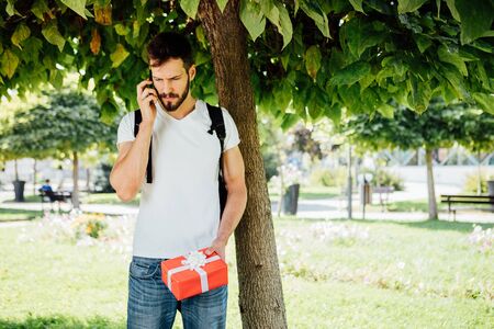handsome man, student, waiting with a red gift next to a treeの写真素材
