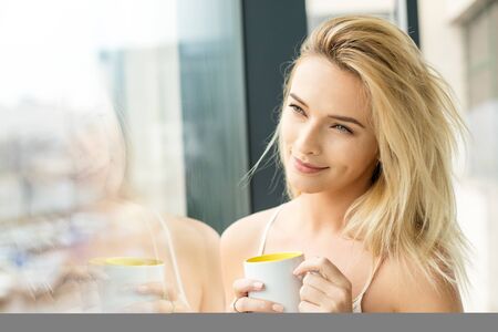 Beautiful blonde woman standing next to a window, wearing night clothes and drinking coffeeの写真素材