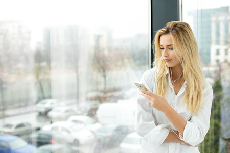 Beautiful blonde woman standing next to a window, wearing men shirt and listening music from mobile phoneの写真素材