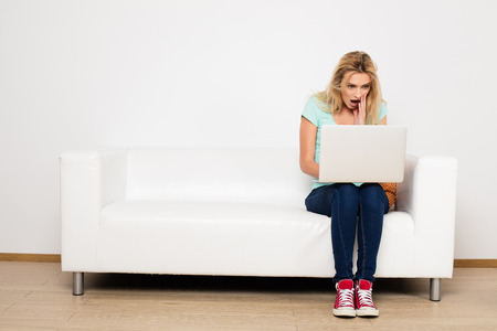 blonde woman sitting on her white sofa with laptop, searching a jobの写真素材