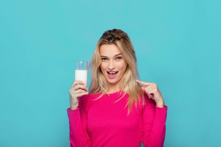 blonde young woman in pink blouse showing a glass of milk on blue backgroundの写真素材