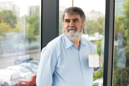 old teacher with gray beard and blue shirt drinking coffee next to a windowの写真素材