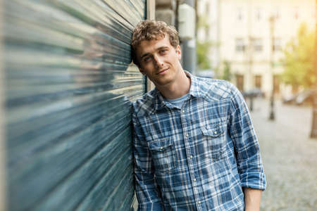 young blond and culry man in checkered shirt standin outside next to a wall