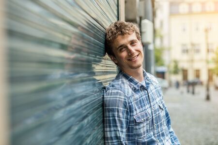 smiling young blond and culry man in checkered shirt standin outside next to a wallの写真素材
