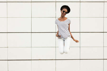 happy modern young black woman with sunglasses, hat and striped shirt  listening music outside and jumpingの写真素材