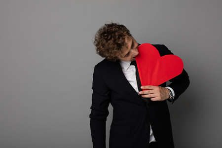 broken heart concept with a curly handsome men biting his red heart on gray backgound