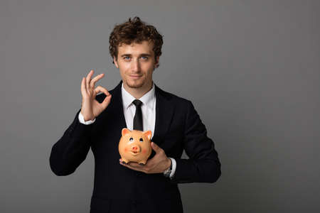 elegant blonde man in suit with blue eyes holding with confidence a piggybankの写真素材