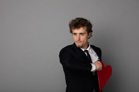 handsome young man in black suit throwing away his red heart, broken heart concept on gray...