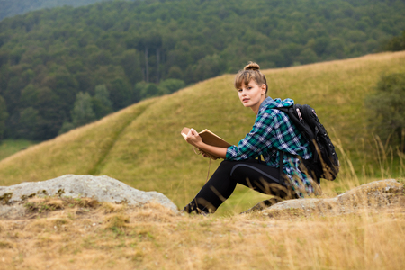 young woman traveling with her backpack in the mountains, sitting on a rock and reading a book or a journalの写真素材