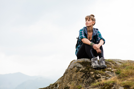young beautiful woman traveling on mountains with backpack on her back, resting on a rock and and watching the viewの写真素材