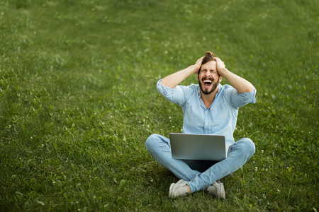 handsome young man lying down in the grass with laptop just gets bad news