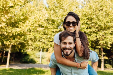 holidays, vacation, love and friendship concept - smiling couple having fun, woman on mens back holding him with love, outside in natureの写真素材
