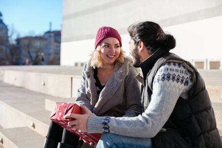 nice joyful couple standing outside on stairs of a building, the boy offering a gift for his girlfriendの写真素材