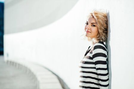 curly blonde woman standing outside and leaning on a wall while looking into cameraの写真素材