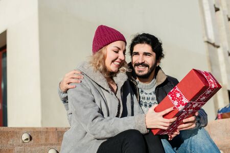 nice joyful couple standing outside on stairs of a building, the boy offering a gift for his girlfriendの写真素材