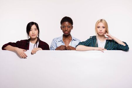 concept of three different ethnicity of young women leaning on a white empty panel smiling and looking to cameraの写真素材