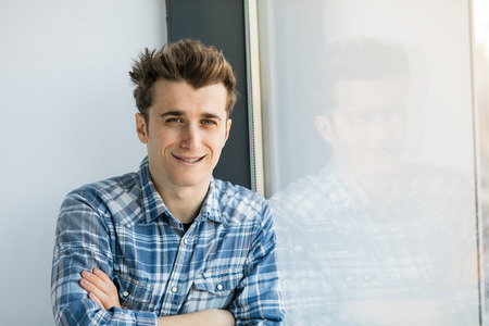 young man with blue eyes in checkeret shirt standing next to a big window with natural light and smiling to cameraの写真素材