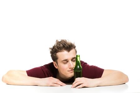 sleepy young man with a bottle of beer sitting at table, isolated on whiteの写真素材