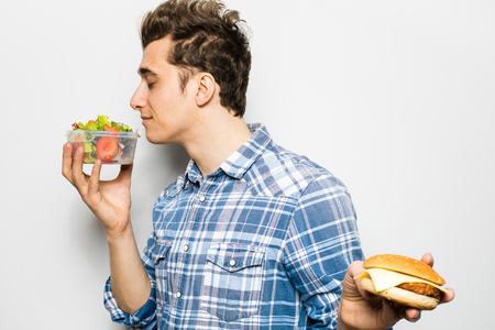 fast food versus healthy food concept with young man holding a salad in one hand and hamburger in the other one, smelling the salad, isolated on whiteの写真素材