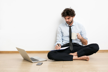A barefoot businessman sitting on the floor against a wall and using a laptop and drinking coffee, working late for his projectの写真素材