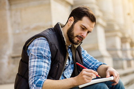 nice student sitting on university stairs outside writing something on his notebookの写真素材