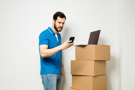 adult man standing next to a stack of carton boxes while using his cellphone and laptop, on white wallの写真素材