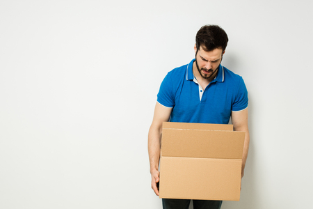 bearded young man in blue shirt with a cardboard box in his arms on a white wallの写真素材