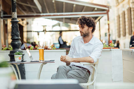 handsome masculine man with curly hair and elegant outfit sitting at a restorant table outside and drinking beerの写真素材
