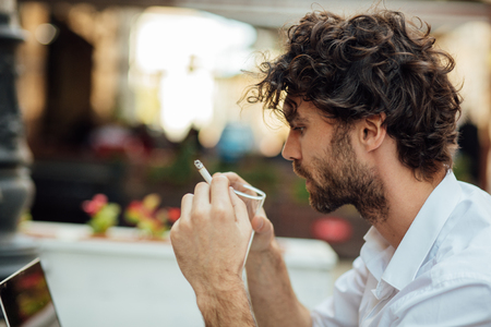 handsome elegant man sitting outside at a restaurant table, working and using laptop while smoking cigarettesの写真素材