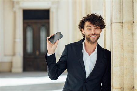 elegant handsome man standing next to columns of an old big building smiling and showing his mobile phoneの写真素材