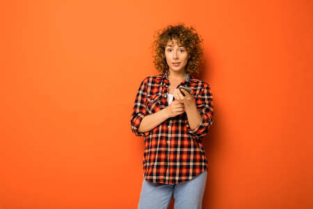 pretty curly woman in a checkered shirt and jeans standing over an orange background with empty space for text next to her with an mobile phone in handsの写真素材