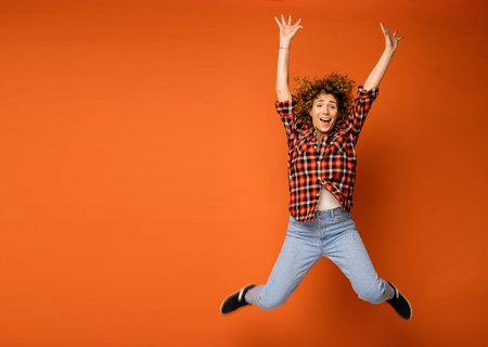 excited curly woman in a checkered shirt and jeans standing over an orange background with empty space for text next to her jumping of happinessの写真素材
