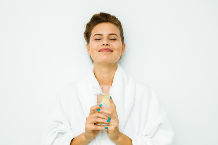 young beautiful woman standing on a white wall in bath towel relaxing with a fresh water with lemonの写真素材