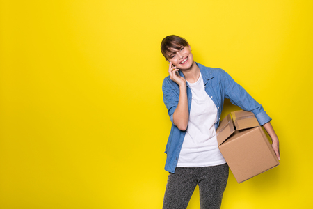 pretty woman standing on yellow background with moving cardboard box and using her cellphoneの写真素材