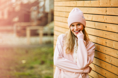 cool hipster young woman in pink hoodie and pink fur cap standing against a wooden wall on the streetの写真素材