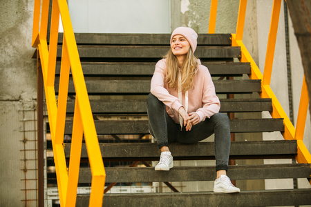 cool hipster young woman in pink hoodie and pink fur cap on some yellow stairs, waiting, playing, being freeの写真素材