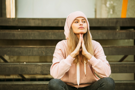cool hipster young woman in pink hoodie and pink fur cap on some yellow stairs, waiting, playing, being freeの写真素材