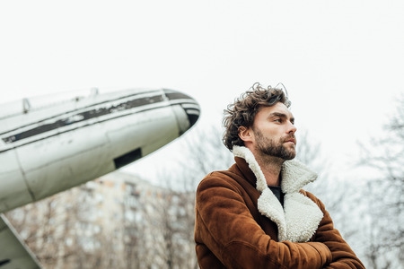 masculine and confident man, in an pilot jacket, standing outside with an old airplane behindの写真素材