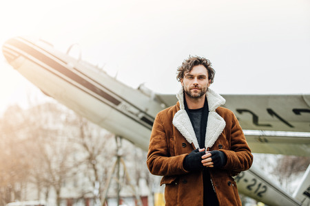 masculine and confident man, in an pilot jacket, standing outside with an old airplane behind