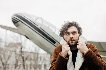 masculine and confident man, in an pilot jacket, standing outside with an old airplane behindの写真素材