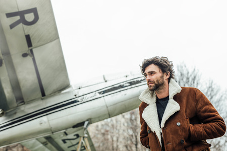 masculine and confident man, in an pilot jacket, standing outside with an old airplane behind