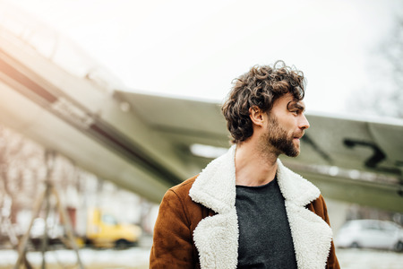 masculine and confident man, in an pilot jacket, standing outside with an old airplane behindの写真素材