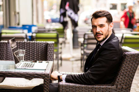 Portrait of handsome successful man with laptop in front looking with confidence at camera, business man having breakfast sitting on beautiful terrace in some old townの写真素材