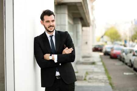 handsome elegant man in suit, businessman, standing outside, waiting for someone, against a white wall with sunglasess, smiling to camera with folded armsの写真素材