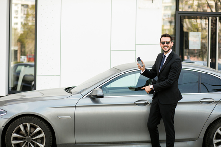 uber driver in elegant suit in front of an luxury car showing his cellphone, outside on the street in front of an office buildingの写真素材