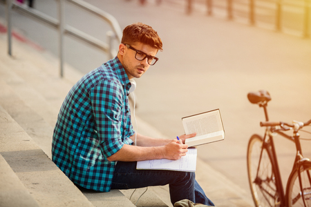 young student with glasses standing on stairs of school or university, looking over his homework, reading a book and taking notesの写真素材