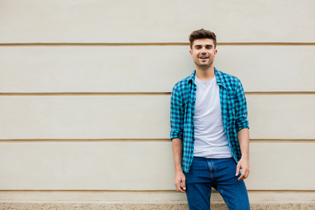 handsome man in checkered shirt smiling standing outside leaning on a wall with free space next to himの写真素材